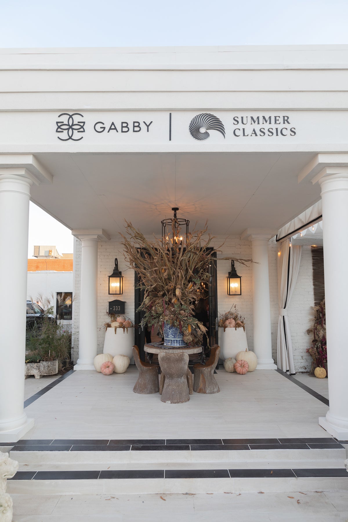 A white outdoor patio or porch area with columns, featuring a central rustic table and chairs arrangement, a large dried floral centerpiece, pumpkins, wall-mounted lanterns, and signage above reading GABBY and SUMMER CLASSICS.
