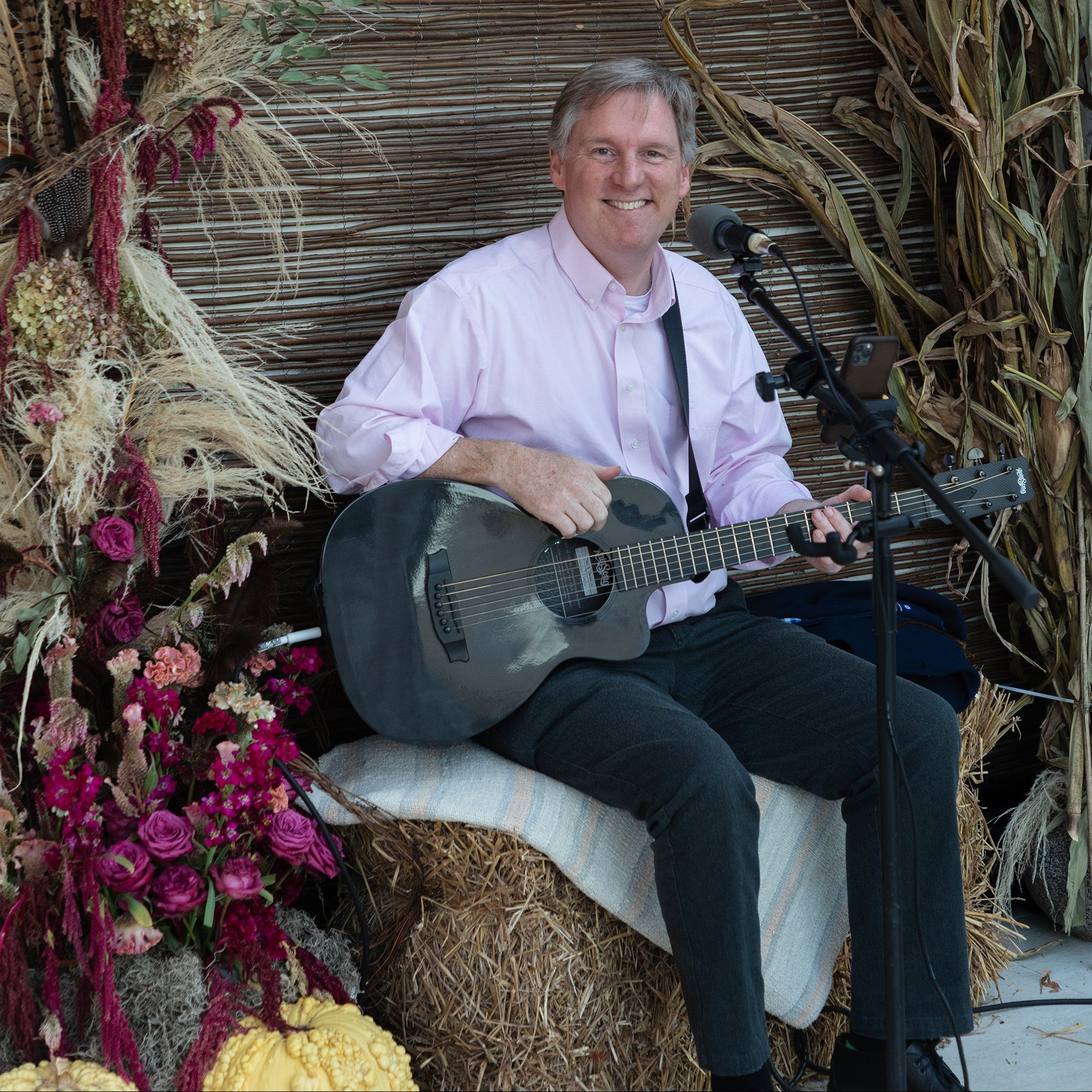 A smiling man sits on a hay bale holding a black acoustic guitar, wearing a light pink dress shirt and dark trousers. He is positioned in front of a rustic brown woven backdrop, framed by a large decorative arch of dried grasses and flowers, with a microphone on a stand.