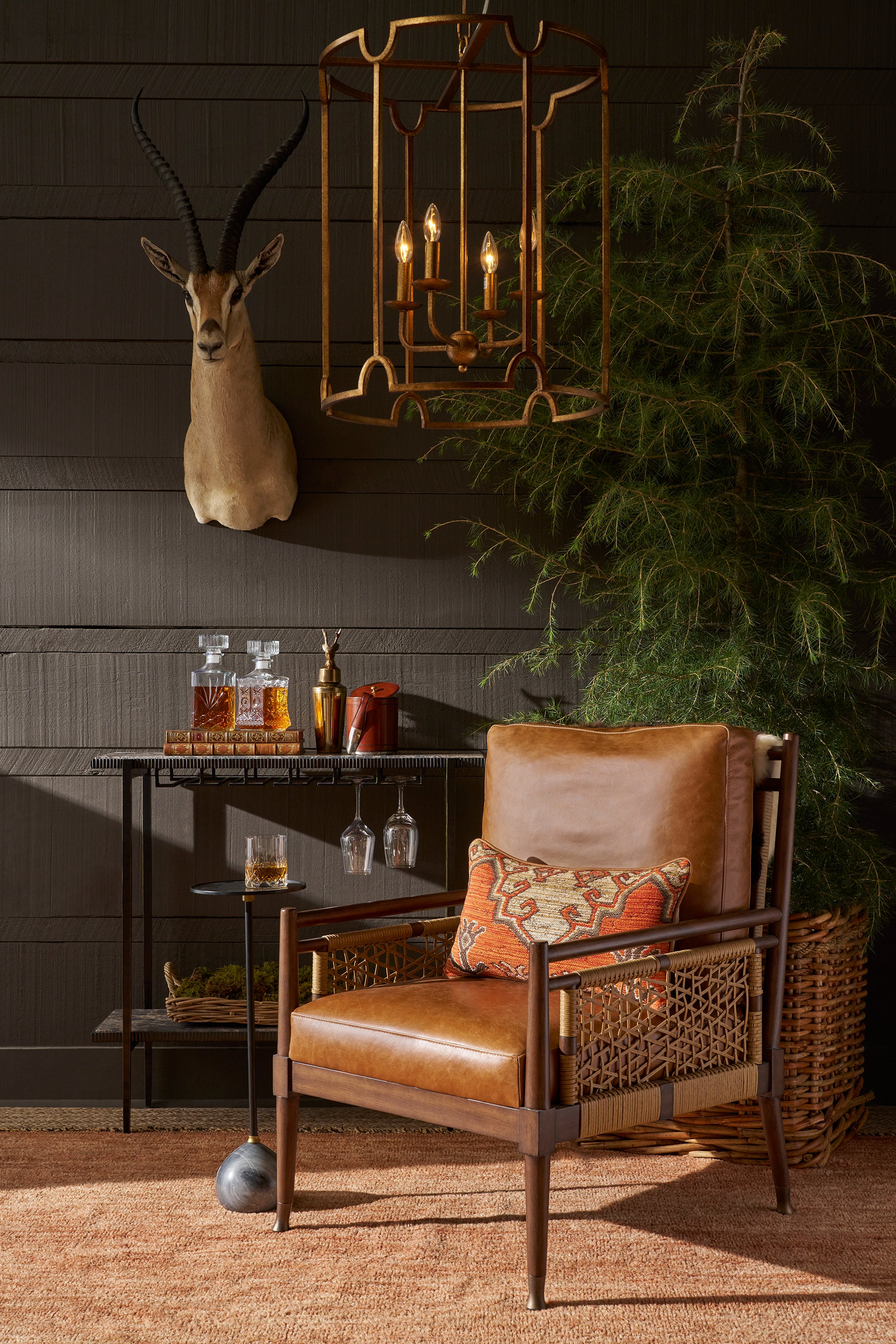 A mid-century modern armchair with a brown leather seat and back, dark wood frame, and woven cane armrests, styled in a rustic room with a patterned accent pillow, a side table, a taxidermy antelope head, and a large green plant.