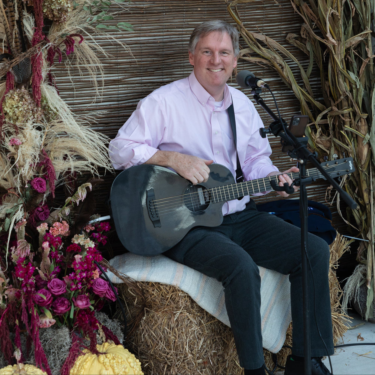 A smiling man sits on a hay bale holding a black acoustic guitar, wearing a light pink dress shirt and dark trousers. He is positioned in front of a rustic brown woven backdrop, framed by a large decorative arch of dried grasses and flowers, with a microphone on a stand.