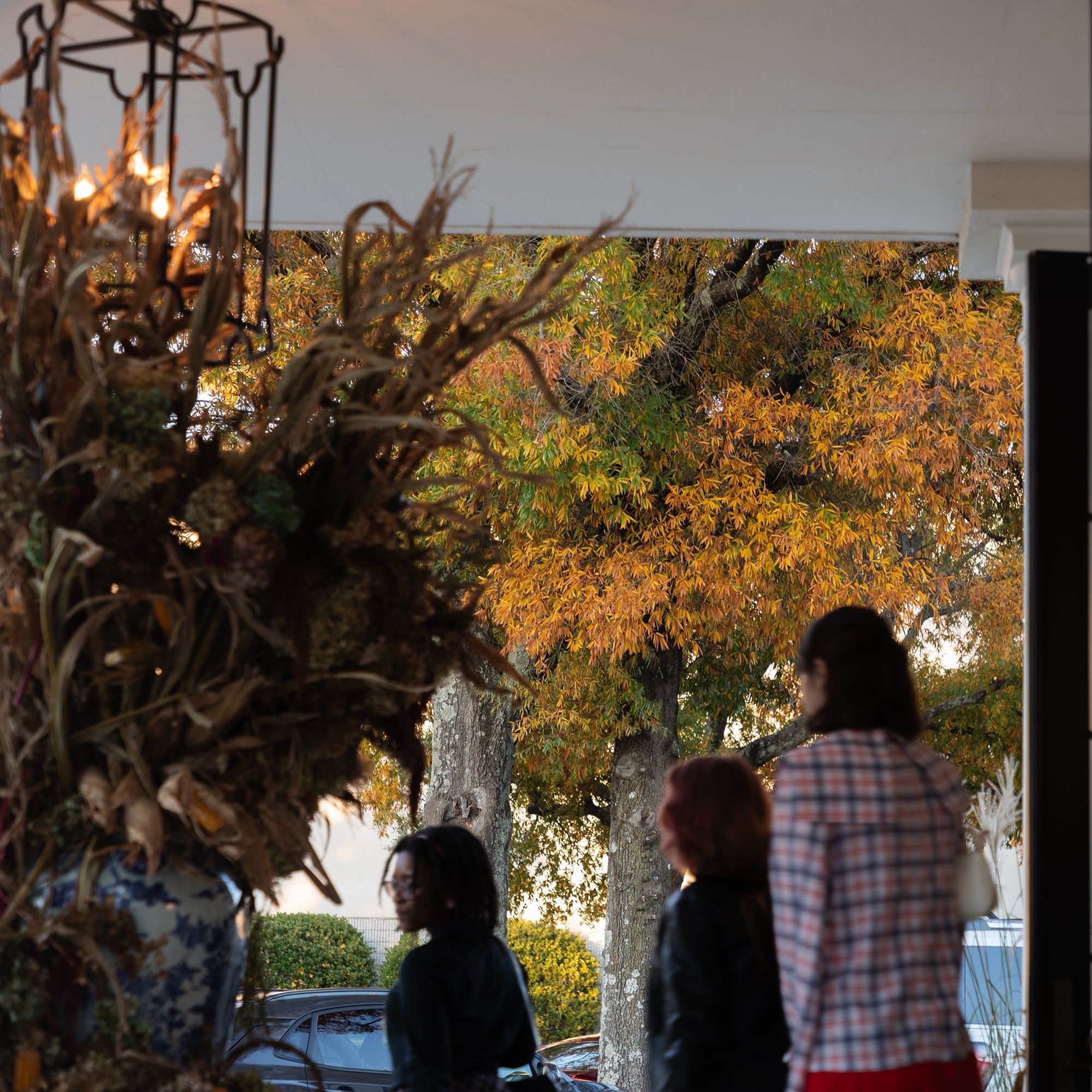 A lifestyle scene at a storefront or porch with three people standing in the doorway, looking out toward trees with autumn orange and yellow leaves. The foreground includes a large decorative dried floral arrangement and a hanging lantern, with a white curtain on the right side.